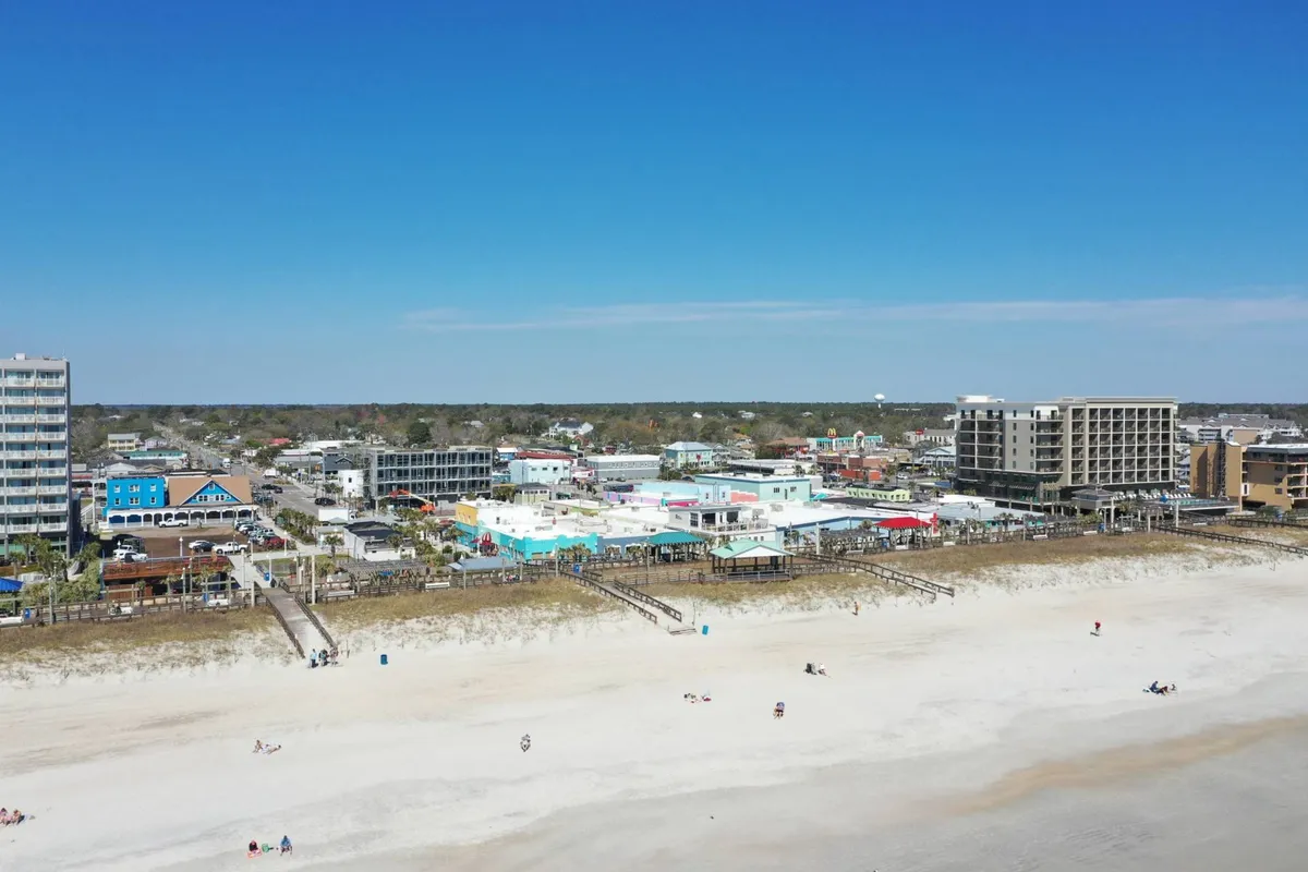 A lively view looking out over the beach, colorful buildings, the bustling boardwalk, and people enjoying the sun, sand, and nearby shops and restaurants. The joyful and relaxed atmosphere is palpable, with families and groups soaking up the vibrant beach scene.