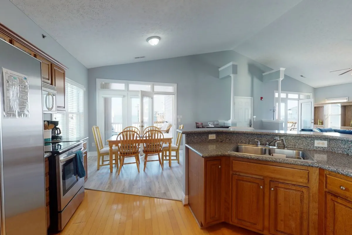 The image showcases a bright, inviting kitchen featuring stainless steel appliances, granite countertops, and a dining table with chairs. Large windows allow ample natural light to fill the space, and the kitchen opens toward an outdoor area, hinting at the possibility of seamless indoor-outdoor dining.