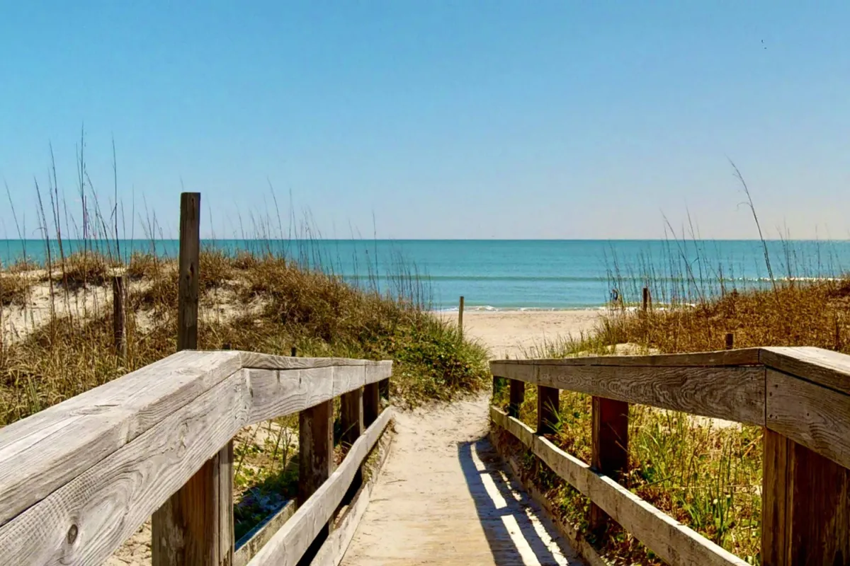 A tranquil wooden walkway leads through the dunes toward the ocean, with the vast sky above and gentle waves in the distance. The serene setting is perfect for morning strolls or sunset walks, and there are seagulls flying overhead, adding to the peaceful coastal atmosphere.