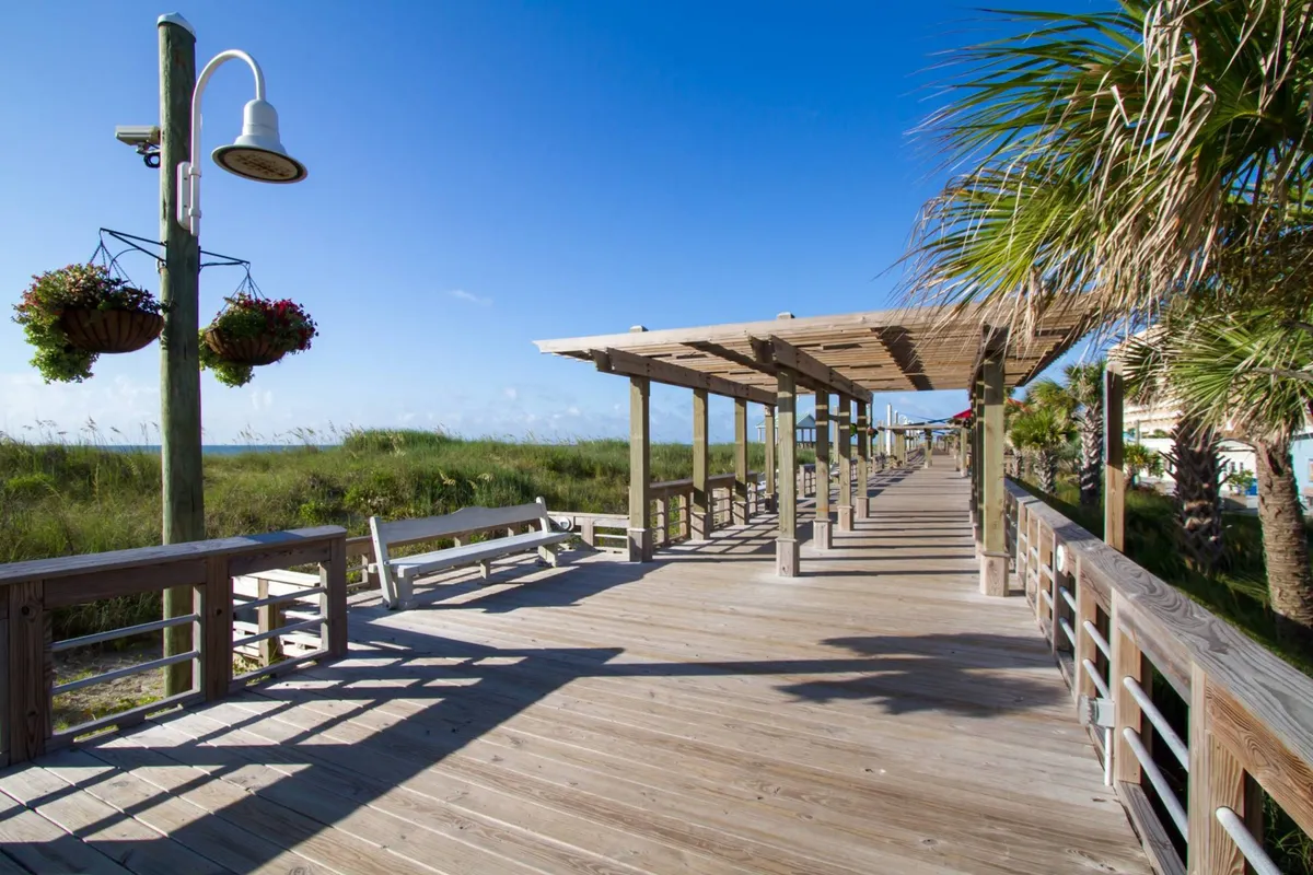 A wooden boardwalk adorned with hanging flower baskets winds through lush greenery and palm trees, inviting visitors to stroll toward the heart of the beach town. The scene is tranquil, yet the carefully maintained walkway suggests easy access to both the ocean and local attractions.