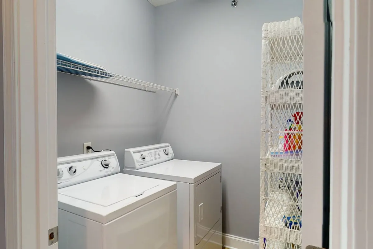 A bright, practical laundry area featuring a washing machine and dryer stacked neatly, with a shelf above holding essential cleaning supplies and a storage rack for organization. The space is clean and easily accessible, emphasizing convenience for guests.