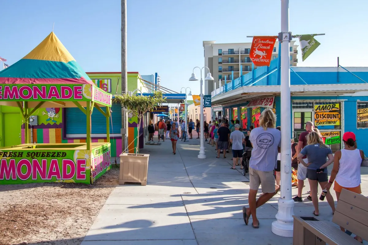 A close-up of the busy Carolina Beach Boardwalk with a brightly colored lemonade stand, bustling storefronts, and people enjoying a lively day out. The focus is on the fun, energetic, and communal atmosphere of the boardwalk's unique local spots.