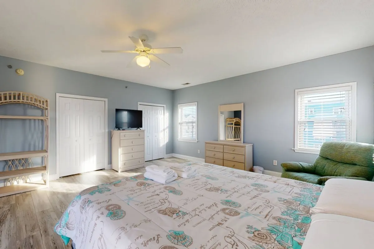 A spacious bedroom featuring a large bed with a decorative quilt, a green armchair in the corner, a flat-screen television, and a wooden dresser. Sunlight streams through the window, creating a warm, tranquil atmosphere ideal for relaxation.