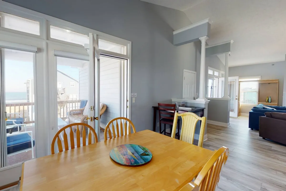 A bright dining area featuring a large table surrounded by chairs, set beside expansive windows that open to the outside deck. The space is bathed in natural light, creating a welcoming environment for family meals, game nights, or relaxed conversations as the sun rises or sets.