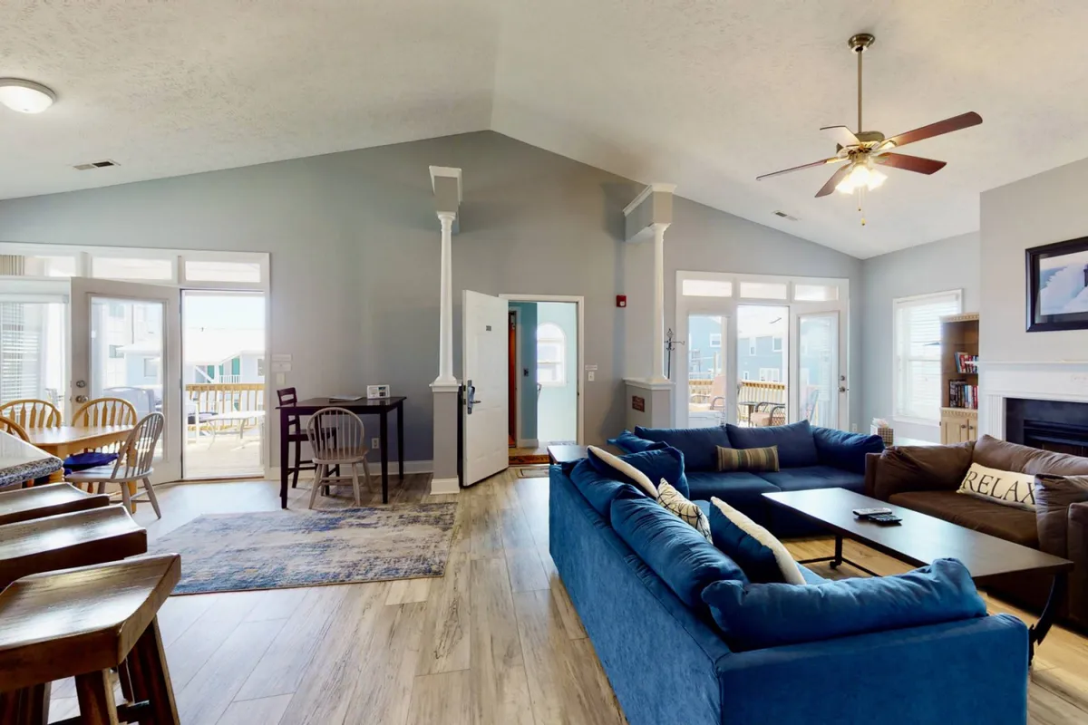 A spacious and inviting living area showcasing a large blue sectional sofa, a wooden dining table, a cozy fireplace, and double doors that allow natural light to fill the space. The room exudes a welcoming, homey atmosphere ideal for relaxing after a day at the beach.