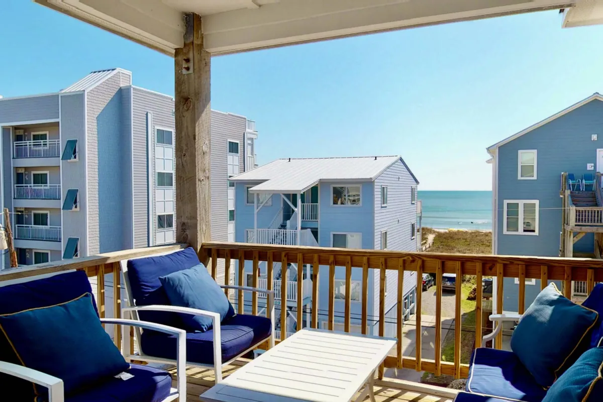 An outdoor deck with wooden balcony railing, comfortable outdoor seating, and a breathtaking, unobstructed view of the blue ocean and sandy beach. The setting invites both morning coffee routines and gatherings to watch the sunrise or sunset, connecting the indoor social spaces with the beauty outside.