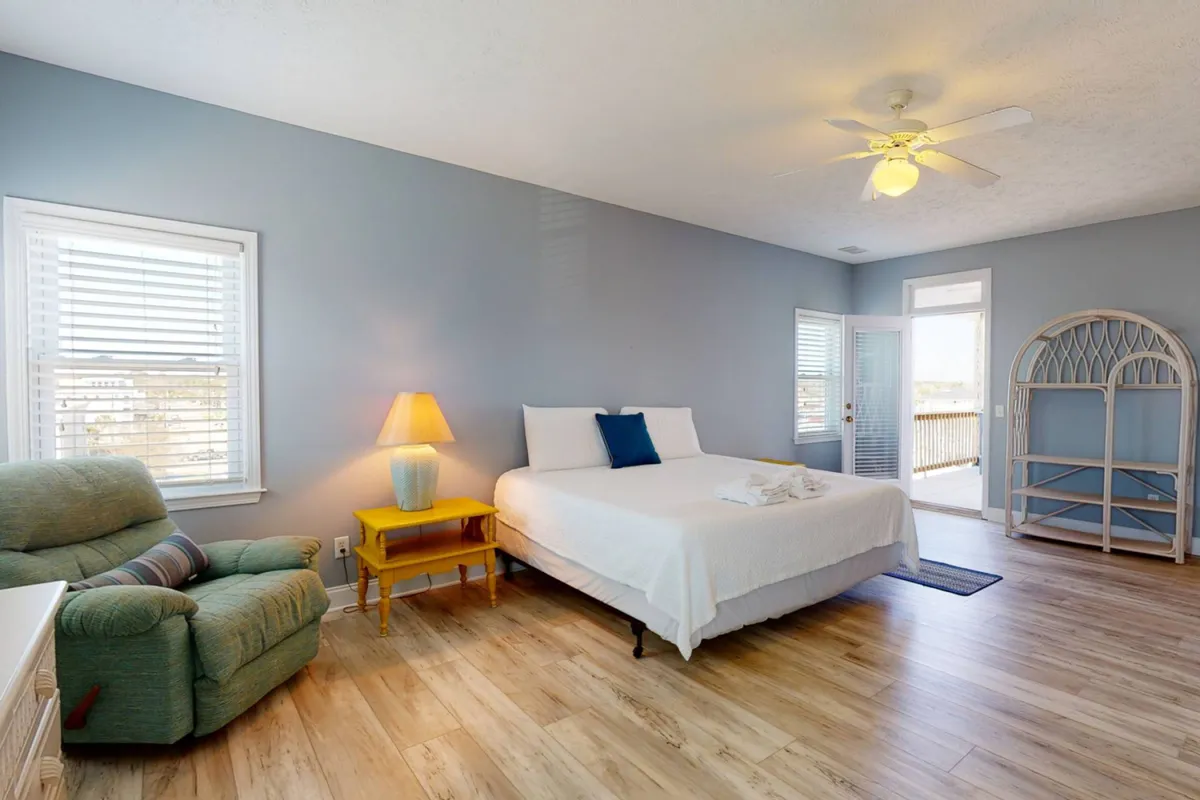 A serene bedroom with a bed dressed in crisp white linens and blue accent pillows. A green armchair and a yellow side table with a lamp provide inviting touches, while large windows fill the room with natural light.