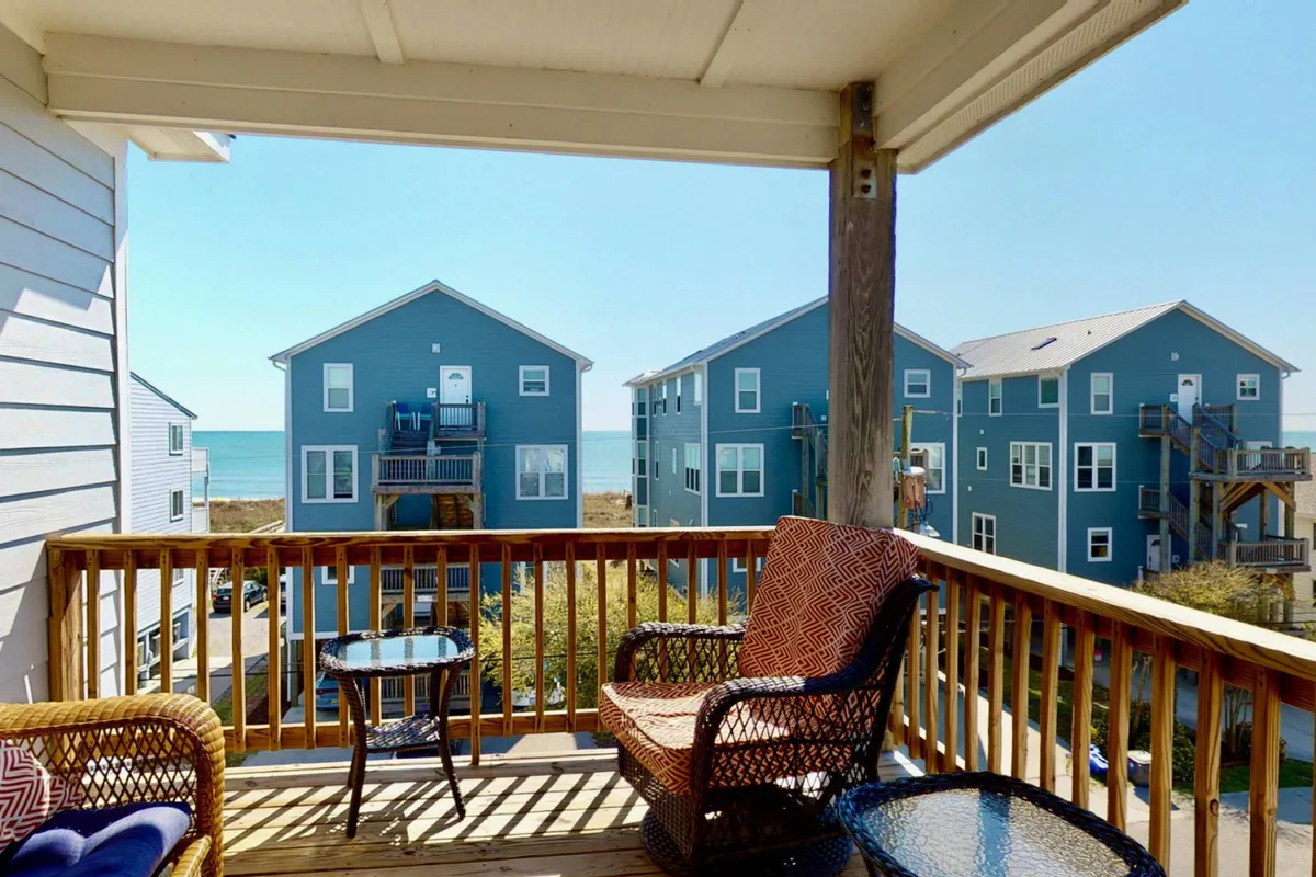 The image showcases a spacious balcony with comfortable seating, overlooking a breathtaking ocean view framed by colorful beachfront buildings. The tranquil scene captures soft sunlight glinting off the waves, suggesting moments of relaxation like savoring morning coffee, reading a book, or gathering with friends at sunset.